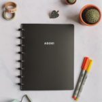 Black notebook with white letters next to assorted items on marble background