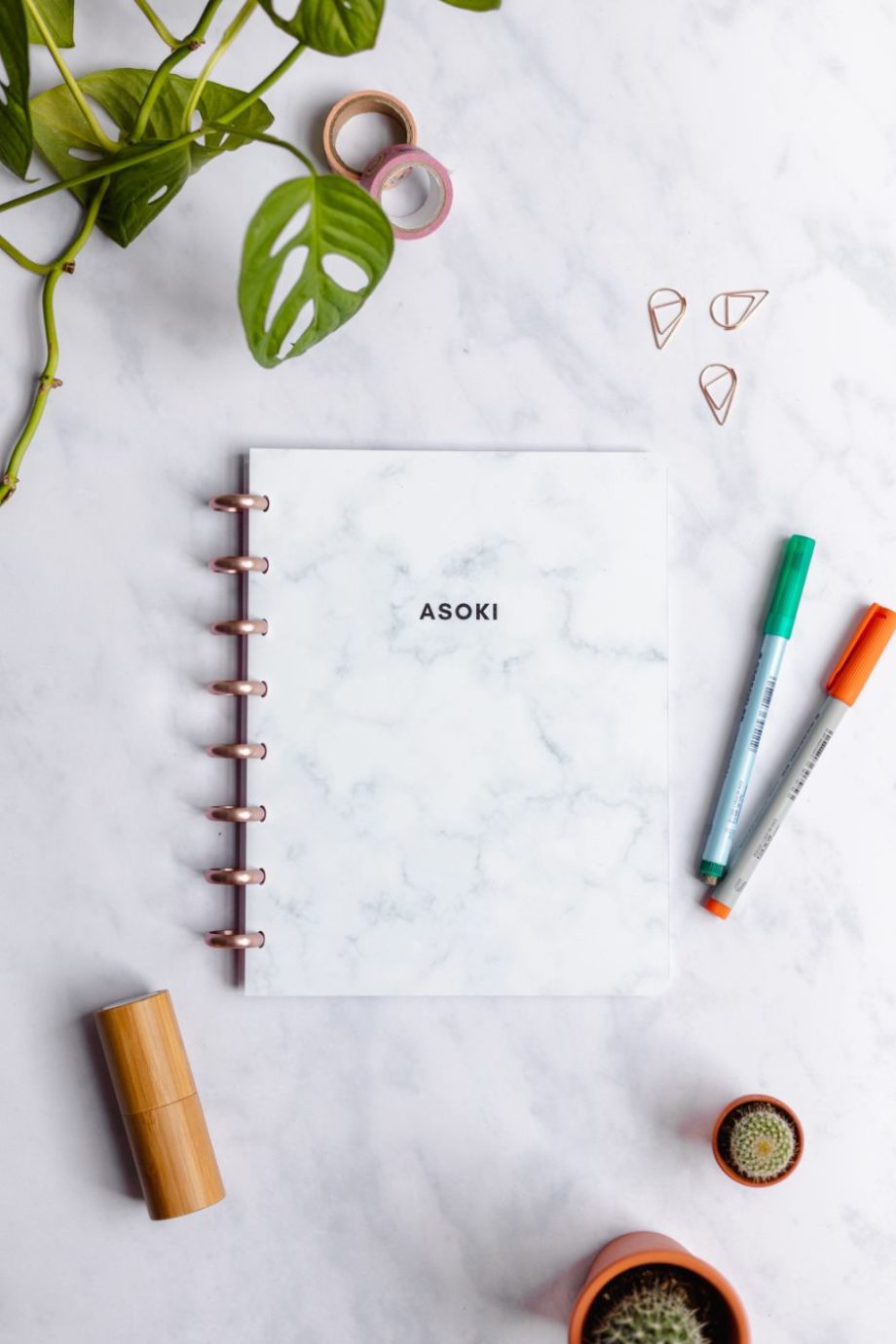 Asoki marble notebook with rose-gold metal discs next to bamboo spray and pens in orange and green on marble background